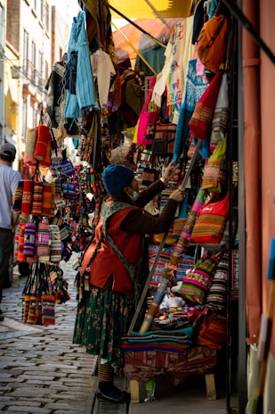 A bustling market scene with a narrow street lined with vibrant textiles and handmade goods. Various colorful fabric items, such as bags and clothes, are displayed on stands and hung around the stall. A person dressed in traditional attire examines the merchandise.