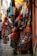 A bustling market scene with a narrow street lined with vibrant textiles and handmade goods. Various colorful fabric items, such as bags and clothes, are displayed on stands and hung around the stall. A person dressed in traditional attire examines the merchandise.