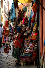 A bustling market scene with a narrow street lined with vibrant textiles and handmade goods. Various colorful fabric items, such as bags and clothes, are displayed on stands and hung around the stall. A person dressed in traditional attire examines the merchandise.
