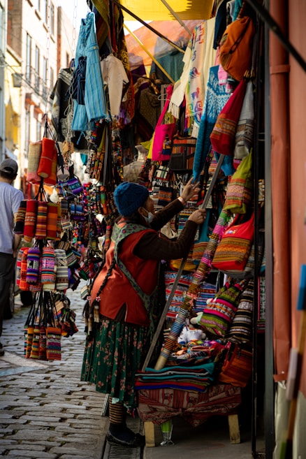 A bustling market scene with a narrow street lined with vibrant textiles and handmade goods. Various colorful fabric items, such as bags and clothes, are displayed on stands and hung around the stall. A person dressed in traditional attire examines the merchandise.