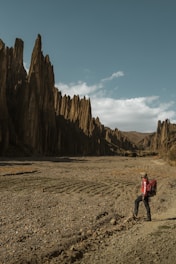 A hiker in outdoor gear stands on a rocky path, gazing at tall, dramatic rock formations under a clear blue sky. The landscape is arid with sparse vegetation, and the rock formations cast long shadows.