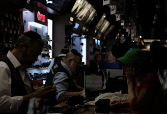 A busy electronics store with multiple counters and customers. Several advertisements and electronics brand logos, such as Canon and SanDisk, are visible on the walls. Customer service representatives in uniform are assisting customers. The setting is dimly lit, giving it an indoor market ambiance.