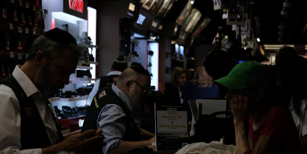 A busy electronics store with multiple counters and customers. Several advertisements and electronics brand logos, such as Canon and SanDisk, are visible on the walls. Customer service representatives in uniform are assisting customers. The setting is dimly lit, giving it an indoor market ambiance.
