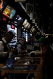 A busy electronics store with multiple counters and customers. Several advertisements and electronics brand logos, such as Canon and SanDisk, are visible on the walls. Customer service representatives in uniform are assisting customers. The setting is dimly lit, giving it an indoor market ambiance.