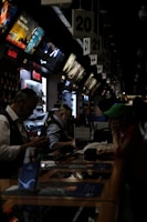 A busy electronics store with multiple counters and customers. Several advertisements and electronics brand logos, such as Canon and SanDisk, are visible on the walls. Customer service representatives in uniform are assisting customers. The setting is dimly lit, giving it an indoor market ambiance.