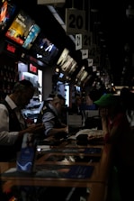 A busy electronics store with multiple counters and customers. Several advertisements and electronics brand logos, such as Canon and SanDisk, are visible on the walls. Customer service representatives in uniform are assisting customers. The setting is dimly lit, giving it an indoor market ambiance.