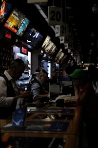 A busy electronics store with multiple counters and customers. Several advertisements and electronics brand logos, such as Canon and SanDisk, are visible on the walls. Customer service representatives in uniform are assisting customers. The setting is dimly lit, giving it an indoor market ambiance.