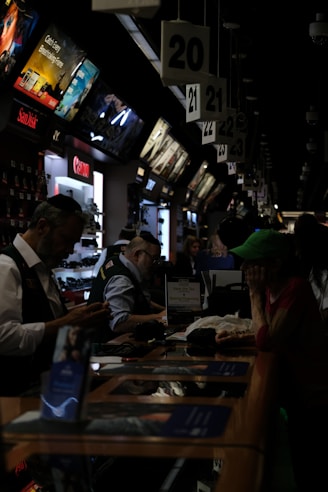 A busy electronics store with multiple counters and customers. Several advertisements and electronics brand logos, such as Canon and SanDisk, are visible on the walls. Customer service representatives in uniform are assisting customers. The setting is dimly lit, giving it an indoor market ambiance.