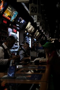 A busy electronics store with multiple counters and customers. Several advertisements and electronics brand logos, such as Canon and SanDisk, are visible on the walls. Customer service representatives in uniform are assisting customers. The setting is dimly lit, giving it an indoor market ambiance.