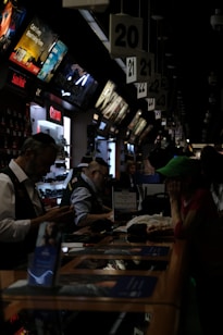 A busy electronics store with multiple counters and customers. Several advertisements and electronics brand logos, such as Canon and SanDisk, are visible on the walls. Customer service representatives in uniform are assisting customers. The setting is dimly lit, giving it an indoor market ambiance.