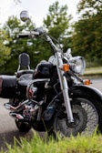 A classic cruiser motorcycle is parked outdoors on a paved surface. It features a shiny chrome finish and black bodywork, with a large front headlight and side mirrors. The background showcases lush green trees and a partly cloudy sky, indicating it is in a natural, serene setting.