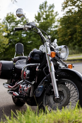 A classic cruiser motorcycle is parked outdoors on a paved surface. It features a shiny chrome finish and black bodywork, with a large front headlight and side mirrors. The background showcases lush green trees and a partly cloudy sky, indicating it is in a natural, serene setting.