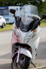 A white Honda scooter is parked on the side of a road with a clear windshield and the brand logo prominently displayed. The background contains blurry images of outdoor vegetation and another vehicle.