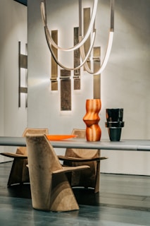 A modern and minimalist dining setup featuring a sleek table and geometrically designed chairs. Above the table hangs an abstract light fixture with curved, elongated elements and warm lighting. Two sculptural vases, one orange and the other black, sit on the table, complemented by an orange tray.