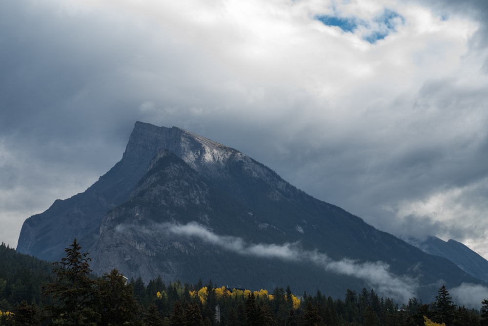 a large mountain with trees in the foreground and clouds in the background