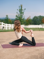 A person is performing a yoga pose on a purple mat in an open outdoor space. The setting is serene with a clear blue sky and lush greenery surrounding the area, including a white wooden fence and trees.