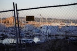A marina filled with various boats docked in the water is visible through a chain-link fence. Several warehouses and buildings are stationed along the waterfront with hills and residential area beyond. A security sign is attached to the fence, and cars are parked near the shore.
