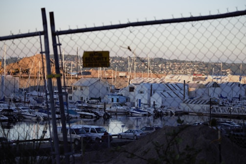 A marina filled with various boats docked in the water is visible through a chain-link fence. Several warehouses and buildings are stationed along the waterfront with hills and residential area beyond. A security sign is attached to the fence, and cars are parked near the shore.