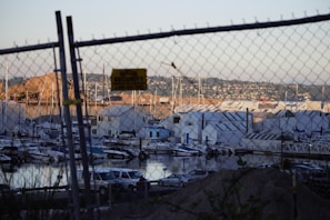 A marina filled with various boats docked in the water is visible through a chain-link fence. Several warehouses and buildings are stationed along the waterfront with hills and residential area beyond. A security sign is attached to the fence, and cars are parked near the shore.