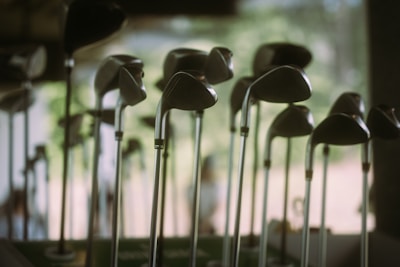 A collection of golf clubs is displayed with their heads positioned at varying heights. The scene appears softly lit, with a blurred background suggesting an indoor setting or a shaded outdoor area. The clubs' metallic surfaces reflect the ambient light, creating a serene and organized atmosphere.