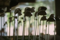 A collection of golf clubs is displayed with their heads positioned at varying heights. The scene appears softly lit, with a blurred background suggesting an indoor setting or a shaded outdoor area. The clubs' metallic surfaces reflect the ambient light, creating a serene and organized atmosphere.