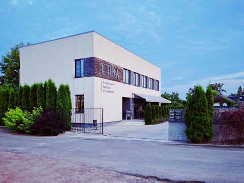 A modern two-story building with a clean, minimalist design and a white facade. The word 'ETIKA' is prominently displayed along with some text in Cyrillic script. The building's exterior is bordered by neatly trimmed tall shrubs, and there is a gated entrance leading to a path made of concrete. The sky above is clear, suggesting a calm day.