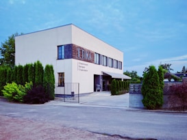 A modern two-story building with a clean, minimalist design and a white facade. The word 'ETIKA' is prominently displayed along with some text in Cyrillic script. The building's exterior is bordered by neatly trimmed tall shrubs, and there is a gated entrance leading to a path made of concrete. The sky above is clear, suggesting a calm day.