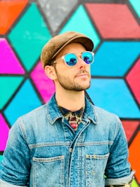 A person wearing a denim jacket, a plaid shirt, and a brown flat cap is looking upwards. They are wearing reflective sunglasses that display a colorful geometric pattern in the lenses. The background consists of a vibrant, multicolored hexagonal pattern, with shades of pink, teal, and blue.