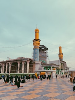 A grand mosque with intricate blue tile work and towering orange minarets is surrounded by scaffolding, possibly undergoing restoration. People, many dressed in dark clothing, fill the courtyard area paved in a patterned design of large square tiles. Some individuals are seated, while others are standing or walking, and one person holds a yellow flag.