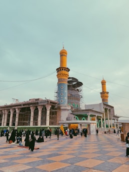 A grand mosque with intricate blue tile work and towering orange minarets is surrounded by scaffolding, possibly undergoing restoration. People, many dressed in dark clothing, fill the courtyard area paved in a patterned design of large square tiles. Some individuals are seated, while others are standing or walking, and one person holds a yellow flag.