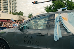A car decorated with the Argentine flag is passing by a crowd of cheering people on a city street. The vehicle has graffiti on the side that reads, 'MIRAS BOBO.' People in the crowd are waving flags and wearing light blue and white, likely supporters or fans celebrating an event. In the background, there are buildings and trees under a partly cloudy sky.
