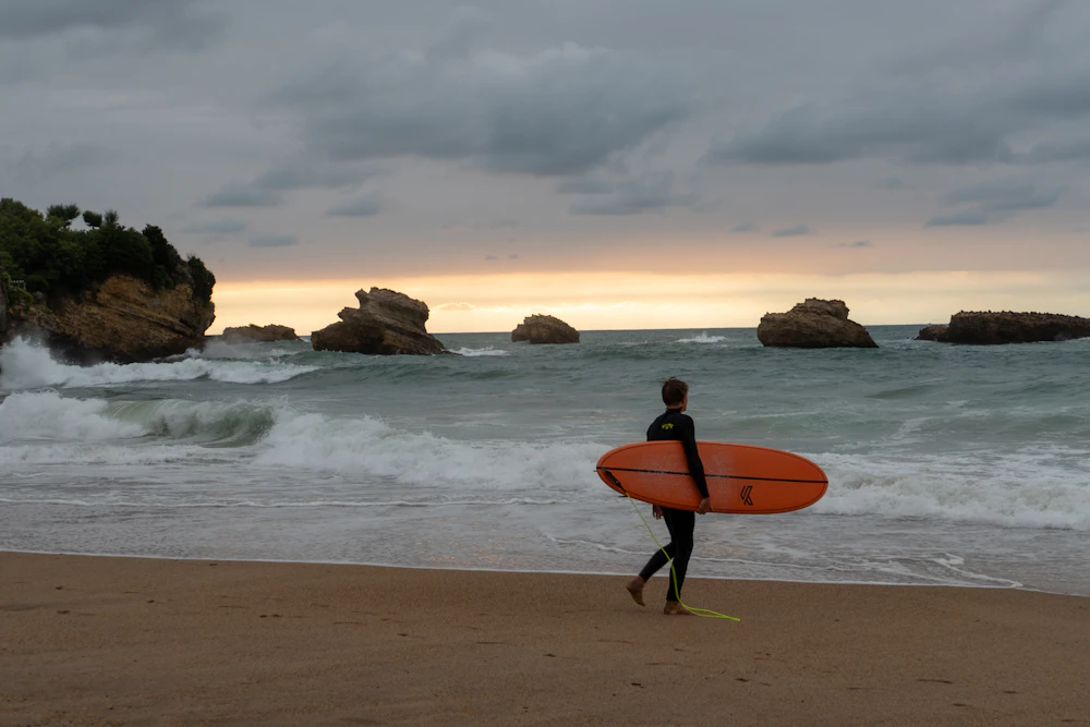 A surfer walking along the beach with a board, rocks offshore