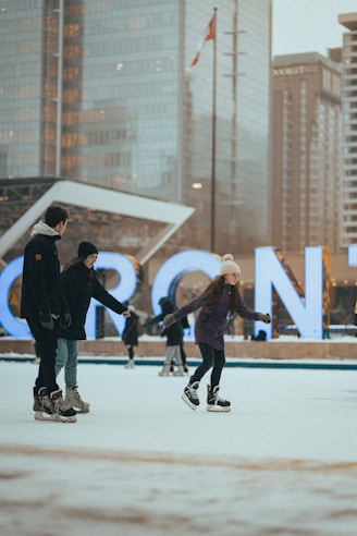 Several people are ice skating in an outdoor rink with tall buildings in the background. The large, illuminated letters spelling out 'Toronto' are visible, and the Canadian flag is flying on a pole.