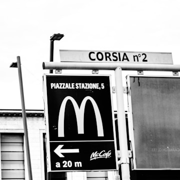 A black and white street sign features the McDonald's logo with directions to a nearby location. Above, another sign displays 'CORSIA n°2'. The image includes parts of buildings and street lights in the background.