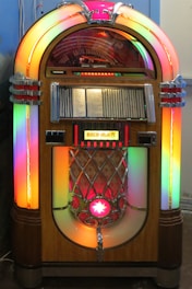 A vintage jukebox with a classic design, featuring colorful neon lights and a wooden exterior. The rounded top houses a visible mechanism for selecting records, while the speaker area below is adorned with metal embellishments and a bright star symbol. The front panel displays song selections.