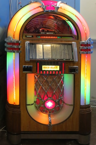 A vintage jukebox with a classic design, featuring colorful neon lights and a wooden exterior. The rounded top houses a visible mechanism for selecting records, while the speaker area below is adorned with metal embellishments and a bright star symbol. The front panel displays song selections.