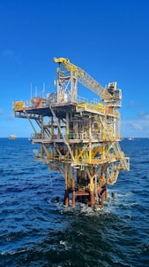 A large offshore oil platform situated in the middle of the ocean with a bright blue sky above. The platform is constructed of metal beams painted in yellow and white with cranes and other equipment visible. The surrounding water is deep blue with gentle waves.