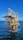 A large offshore oil platform situated in the middle of the ocean with a bright blue sky above. The platform is constructed of metal beams painted in yellow and white with cranes and other equipment visible. The surrounding water is deep blue with gentle waves.