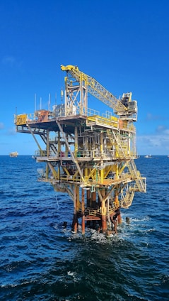 A large offshore oil platform situated in the middle of the ocean with a bright blue sky above. The platform is constructed of metal beams painted in yellow and white with cranes and other equipment visible. The surrounding water is deep blue with gentle waves.