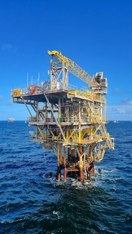A large offshore oil platform situated in the middle of the ocean with a bright blue sky above. The platform is constructed of metal beams painted in yellow and white with cranes and other equipment visible. The surrounding water is deep blue with gentle waves.