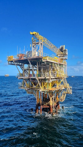A large offshore oil platform situated in the middle of the ocean with a bright blue sky above. The platform is constructed of metal beams painted in yellow and white with cranes and other equipment visible. The surrounding water is deep blue with gentle waves.