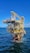 A large offshore oil platform situated in the middle of the ocean with a bright blue sky above. The platform is constructed of metal beams painted in yellow and white with cranes and other equipment visible. The surrounding water is deep blue with gentle waves.