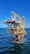 A large offshore oil platform situated in the middle of the ocean with a bright blue sky above. The platform is constructed of metal beams painted in yellow and white with cranes and other equipment visible. The surrounding water is deep blue with gentle waves.