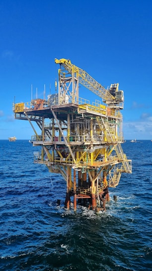 A large offshore oil platform situated in the middle of the ocean with a bright blue sky above. The platform is constructed of metal beams painted in yellow and white with cranes and other equipment visible. The surrounding water is deep blue with gentle waves.