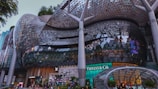 A modern architectural structure with a large glass and steel facade featuring intricate patterns. The building includes well-known luxury brand stores. There are people walking and a landscaped exterior with greenery.