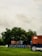 A collection of shipping containers stacked in a grassy field, with a backdrop of dense green trees. The containers bear different logos and colors including red and blue. The sky is overcast with gray clouds, suggesting a moody atmosphere.