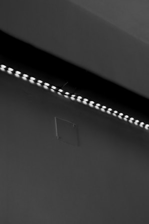 A row of small LED lights emitting a soft glow along the edge of a dark, flat surface. The surface appears smooth with a slight texture, possibly a wall or ceiling. The overall setting is dimly lit, creating a stark contrast between the lights and the surrounding darker area.