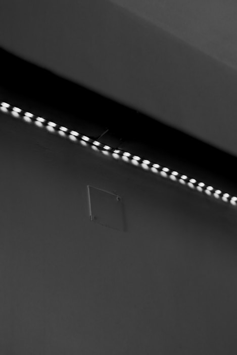 A row of small LED lights emitting a soft glow along the edge of a dark, flat surface. The surface appears smooth with a slight texture, possibly a wall or ceiling. The overall setting is dimly lit, creating a stark contrast between the lights and the surrounding darker area.