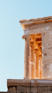 A section of an ancient Greek temple with tall, ornate columns and intricate stone carvings, highlighted by warm sunlight against a clear blue sky.