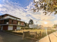 A modern building with large glass windows and wooden accents stands near a construction site. The sun is setting, casting a warm glow over the scene. The sky is filled with scattered clouds, and a construction crane is visible in the background. Trees and a fence line the area, contributing to a serene and peaceful atmosphere.